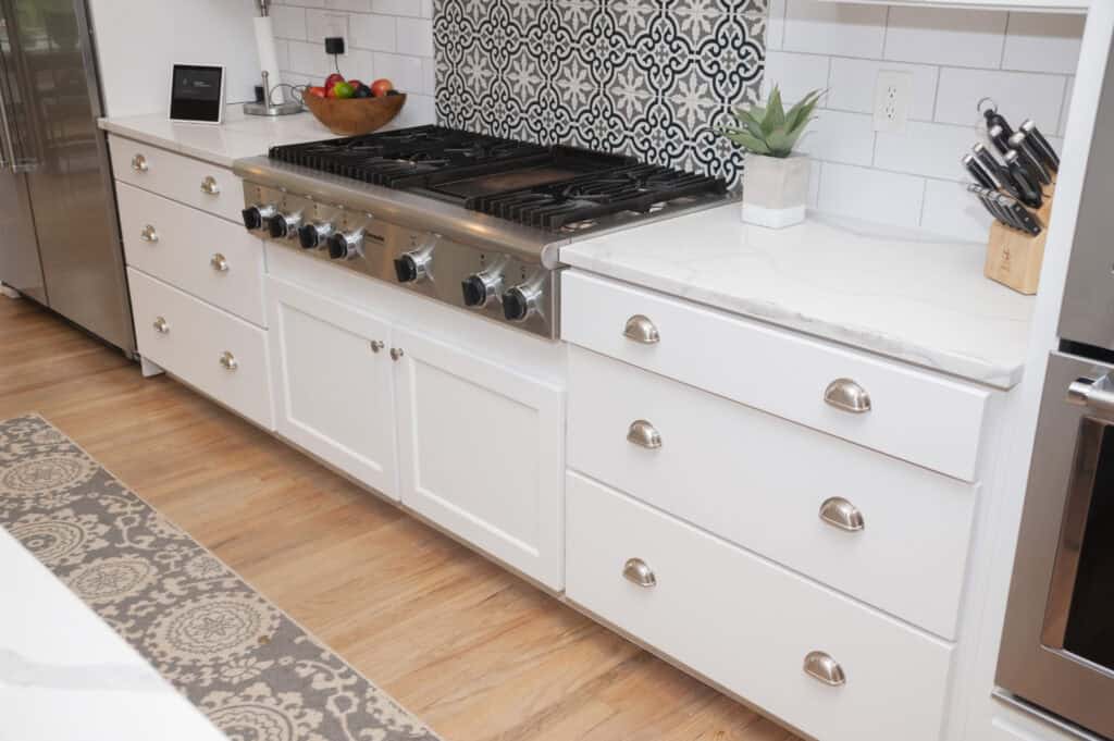 A modern kitchen features a white marble countertop, a stainless steel gas stove with four burners, white cabinets with silver handles, a patterned tile backsplash, a knife block, a fruit bowl, and a small potted plant.