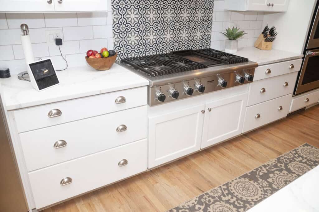 White kitchen cabinets with silver handles, a stainless steel gas stove, patterned black-and-white backsplash, a bowl of fruit, a digital display, a plant, and knives on a wooden floor with a rug.