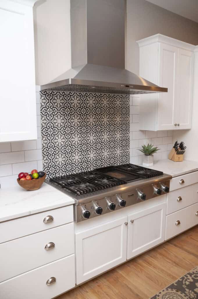 A modern kitchen features a stainless steel gas stove with a patterned black and white tile backsplash, white cabinets, a wooden bowl of fruit, a small potted plant, and a knife block on a white countertop.