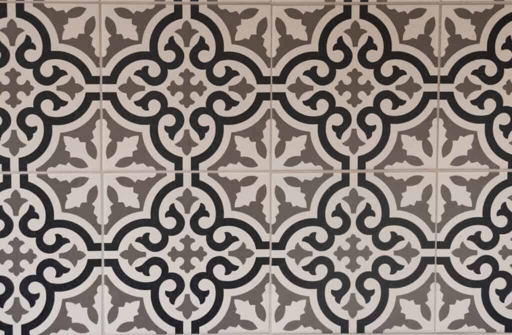 A close-up of decorative ceramic tiles featuring a repeating geometric and floral pattern in black, white, and beige tones. The design appears symmetrical and vintage in style.