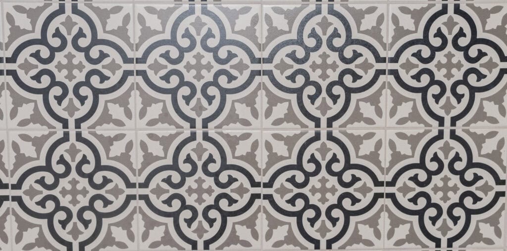 A close-up of decorative ceramic tiles featuring a repeating geometric and floral pattern in shades of gray, white, and black. The intricate design creates a symmetrical, vintage-inspired look.