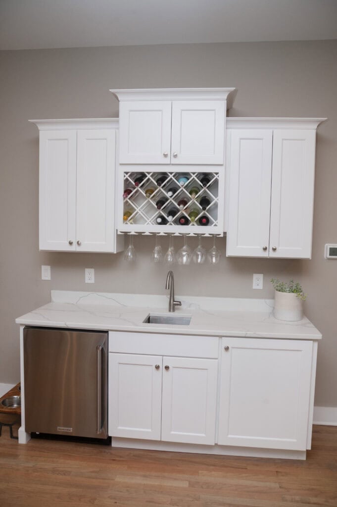 A modern kitchen with white cabinets, a built-in wine rack, hanging wine glasses, a stainless steel dishwasher, a small sink, and a potted plant on the countertop. The floor is wood, and the wall is beige.
