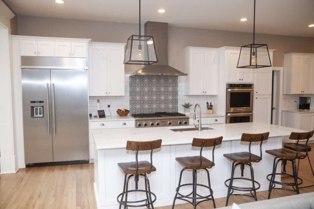 Modern kitchen with white cabinets, stainless steel appliances, a patterned tile backsplash, marble island countertop, four wooden barstools, and pendant lights hanging above the island.