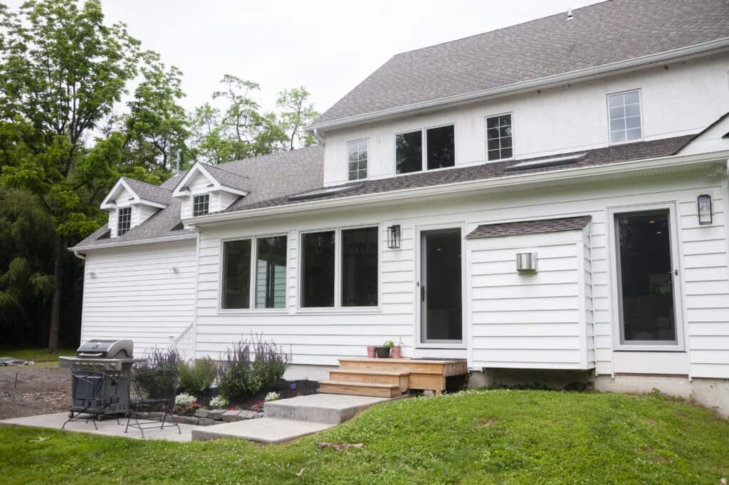 A white two-story house with large windows and a small wooden porch. A grill and flower bed sit beside the porch, and the house is surrounded by green grass and tall trees.
