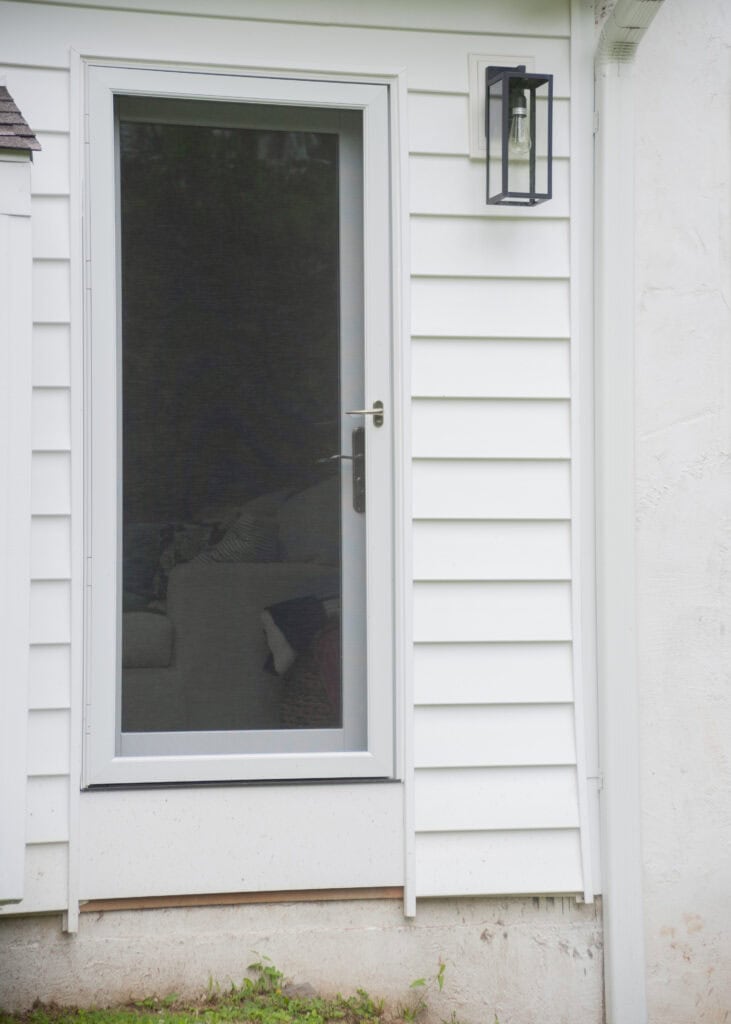 A glass storm door with a metal handle is set in white siding. A black outdoor lantern is mounted above the door, and part of a couch and pillows are visible through the glass. Grass grows at the base of the wall.