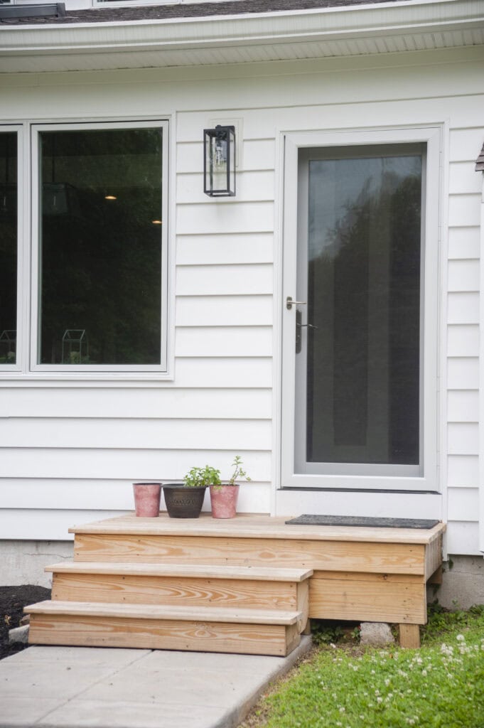 A small wooden porch with three steps leads to a glass door on a white house. Three potted plants sit on the steps. A large window and an outdoor light fixture are next to the door.