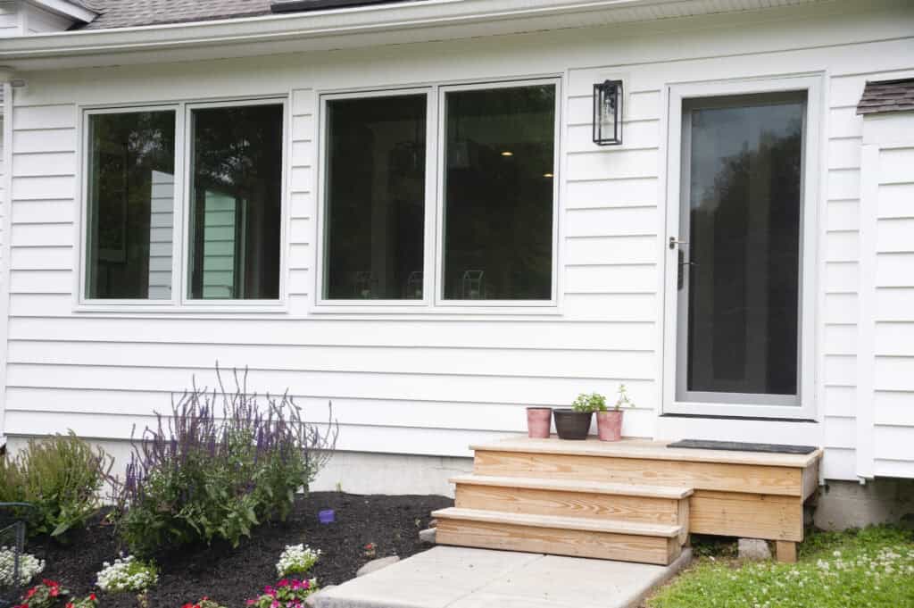 A set of wooden steps leads to a glass storm door on a white house. Three small potted plants sit on the steps, with flower beds and blooming plants bordering the walkway.
