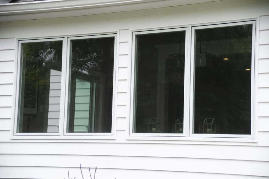 Three large rectangular windows set in a white paneled house exterior reflect greenery and trees from outside. The windows are side by side, with white frames and slightly open.