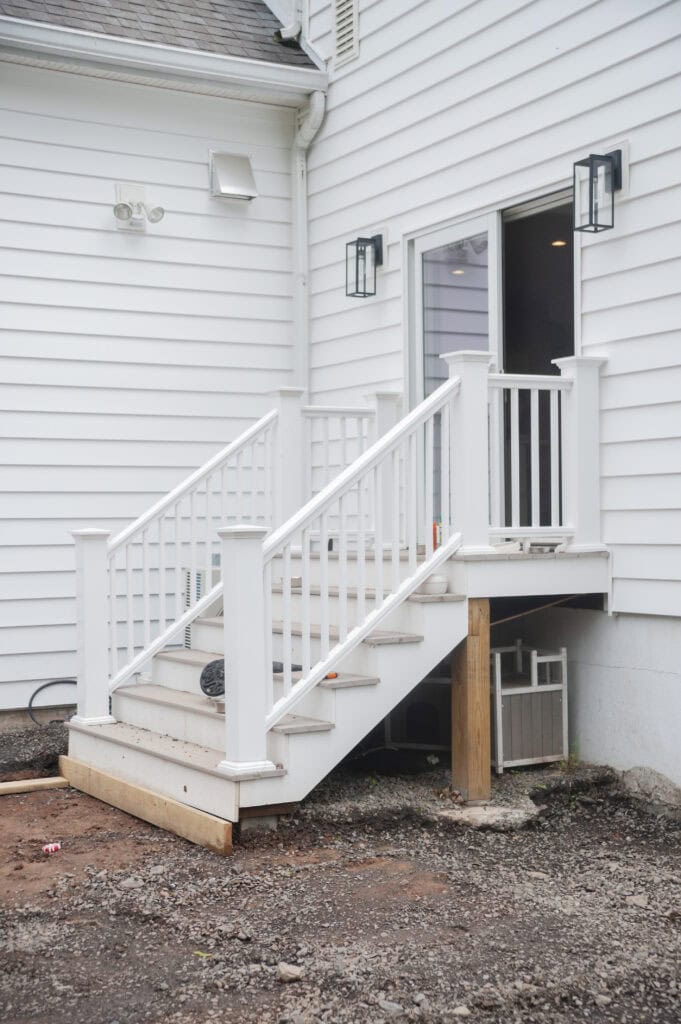 A small white staircase with railing leads from a sliding glass door to the ground outside a white house. The stairs are unfinished at the bottom, with exposed lumber and dirt around the base.