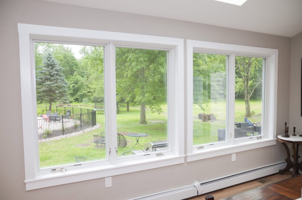 Large white-framed windows provide a clear view of a green backyard with trees, patio furniture, and a firepit. The interior walls are light beige with wood flooring and white trim.