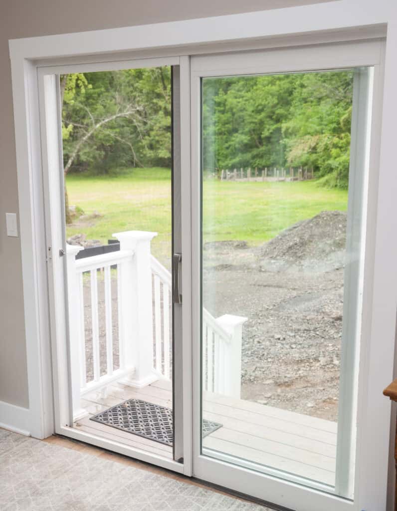 A glass sliding door, partially open, leads from a carpeted indoor space to a small outdoor porch with white railings. Beyond the porch is a green yard with trees and a pile of dirt.