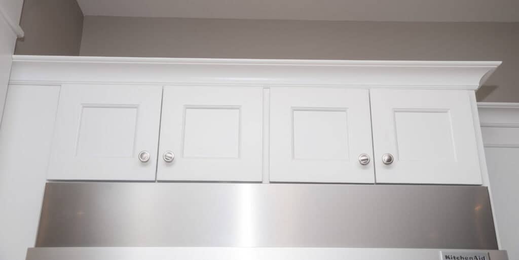 White kitchen cabinets with four doors, each having a silver knob, are mounted above a stainless steel appliance against a light beige wall.