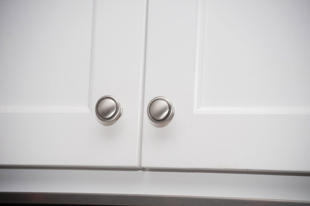 Close-up of two white cabinet doors with brushed metal round knobs, showing a slight gap between the doors. The edges of the doors and knobs are clearly visible.