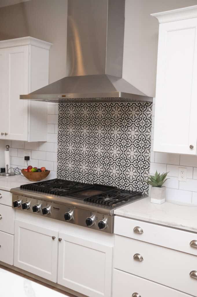 Modern kitchen with white cabinets, a stainless steel stove and range hood, decorative black and white patterned backsplash, a bowl of fruit, and a small potted plant on a white countertop.