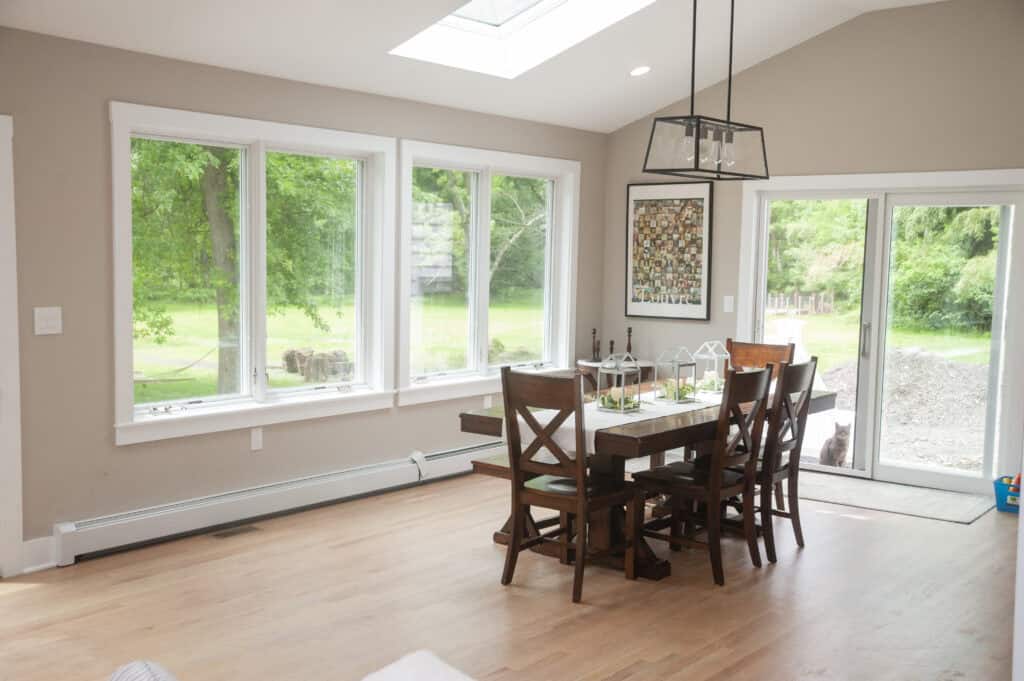 A bright dining area with large windows and glass doors showing a green yard. There is a wooden dining table with six chairs and decorative items, light walls, and light wood floors. Natural light fills the room.