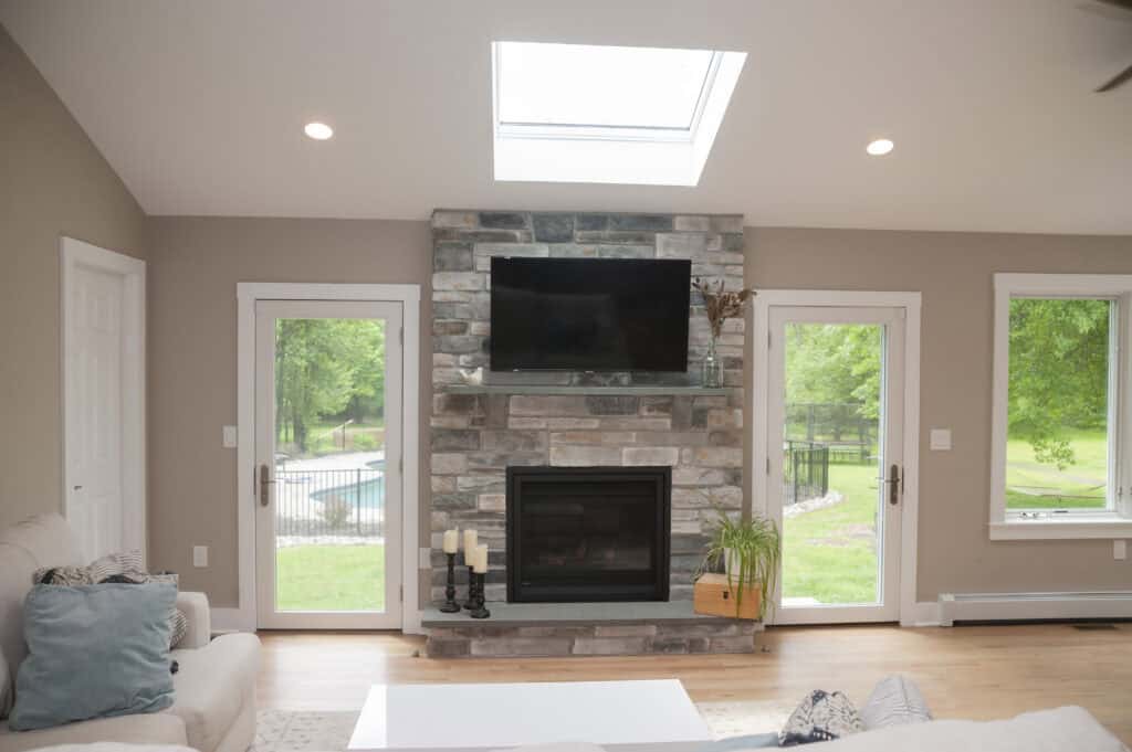 A modern living room with a stone fireplace and mounted TV, flanked by two glass doors showing a green backyard and pool. A skylight above lets in natural light, and light-colored furniture decorates the space.