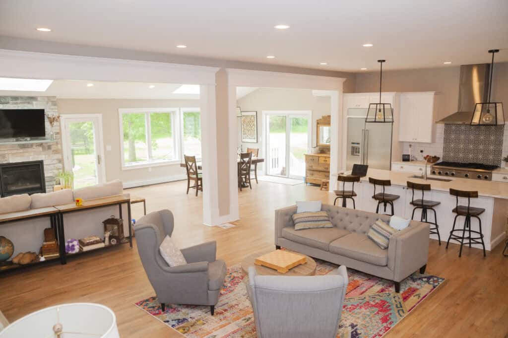 An open-concept living area with gray armchairs, a tufted sofa, a colorful rug, a kitchen with white cabinets, bar stools, and a dining area by large windows overlooking a green yard.