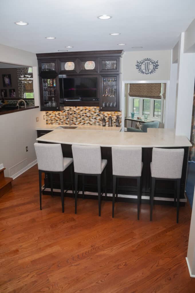 A modern home bar with three upholstered barstools, a light countertop, dark cabinetry, a backsplash with neutral tiles, glass shelves, and a mounted TV above the counter. Wood floors and stairs are visible.