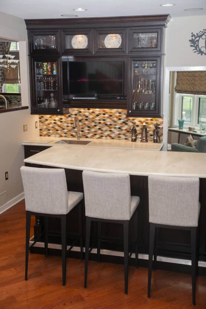 Home bar area with dark wood cabinets, a mounted TV, glass shelving, a sink, and a colorful tiled backsplash. Three light gray upholstered barstools sit at a light-colored countertop on hardwood flooring.