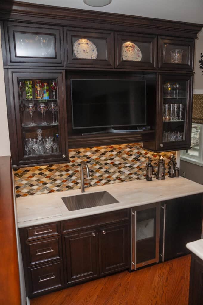 A modern wet bar features dark wood cabinets, a marble countertop with a sink, glassware, a wine fridge, a colorful mosaic backsplash, and a wall-mounted TV above the counter.