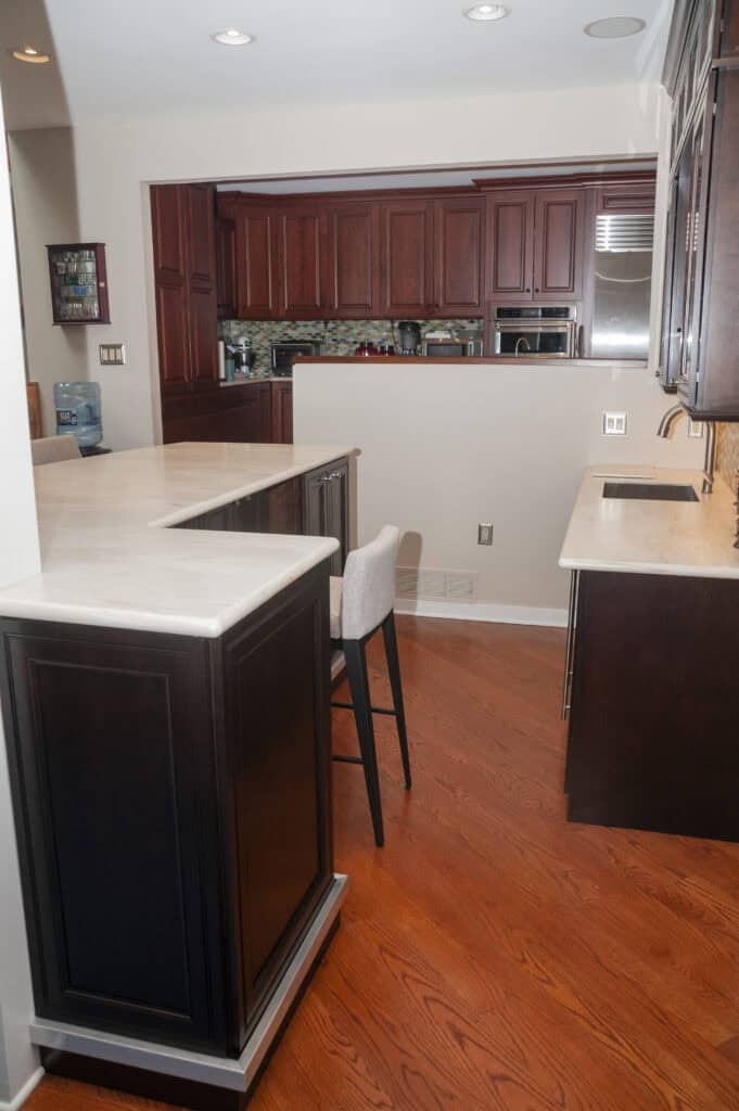 A modern kitchen with dark wood cabinets, white countertops, hardwood floors, and a single upholstered bar stool at an L-shaped counter. The space is well-lit with recessed ceiling lights.