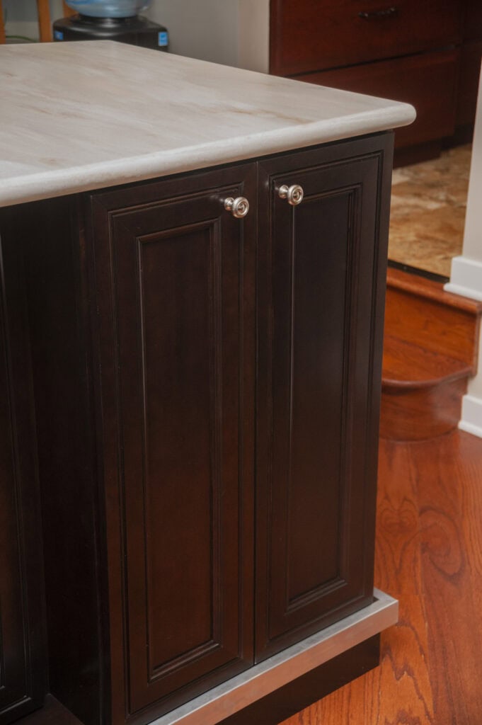 Dark wood kitchen cabinets with silver knobs beneath a light-colored marble countertop, set on a polished wooden floor, next to a step leading to another room.