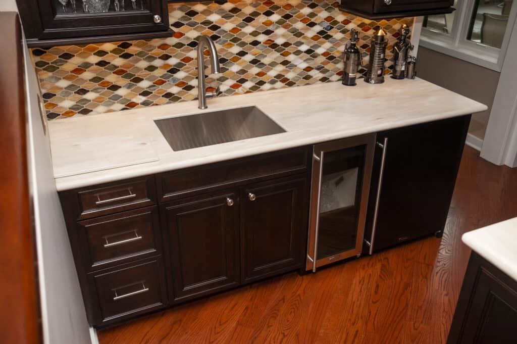Modern kitchen with dark wood cabinets, a stainless steel sink and faucet, a small wine fridge, light countertop, and a colorful diamond-patterned tile backsplash. The floor is a warm, polished wood.