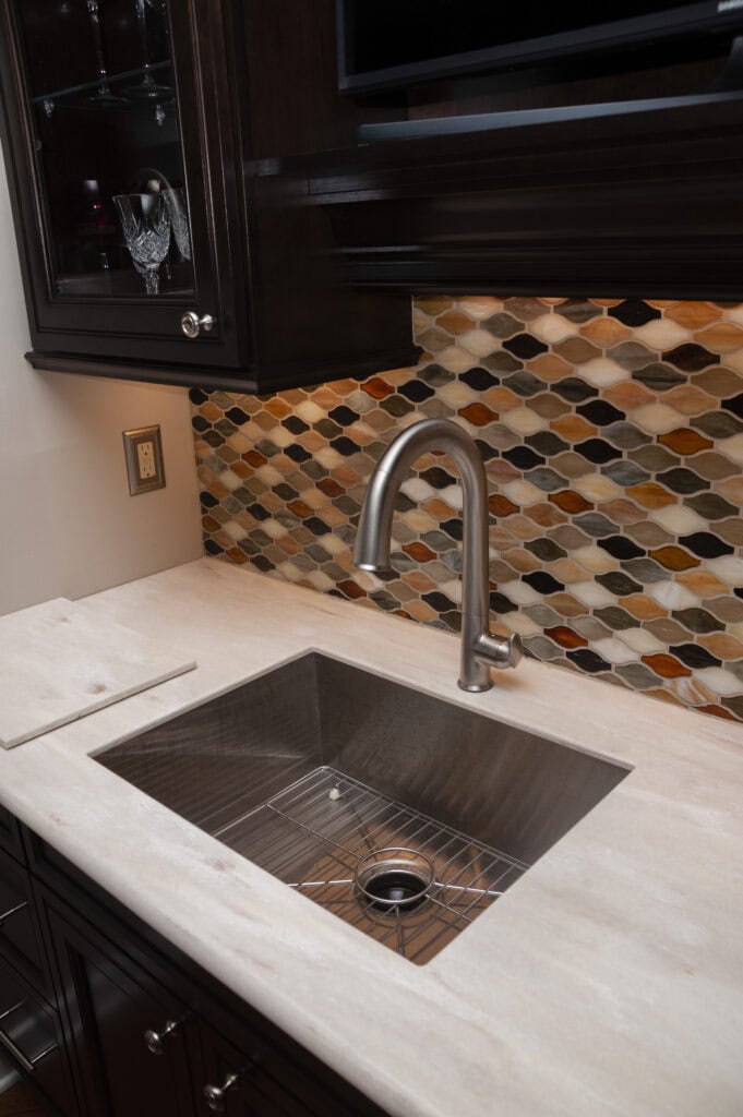 A modern kitchen sink with a stainless steel faucet, set in a light countertop, and a colorful, geometric tile backsplash in shades of brown, orange, and cream. Dark cabinets are visible above and below the sink.