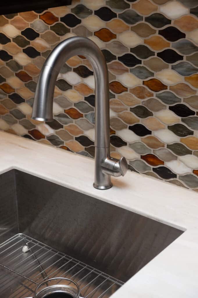 A modern stainless steel kitchen faucet over a deep metal sink, set against a colorful, geometric-patterned tile backsplash in shades of brown, orange, and gray.