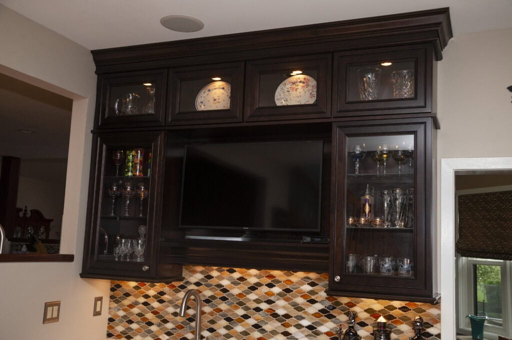 Dark wood kitchen cabinets above a sink display decorative plates and glassware. A flat-screen TV is set in the center, with a colorful mosaic tile backsplash below. Spotlights illuminate the shelves.