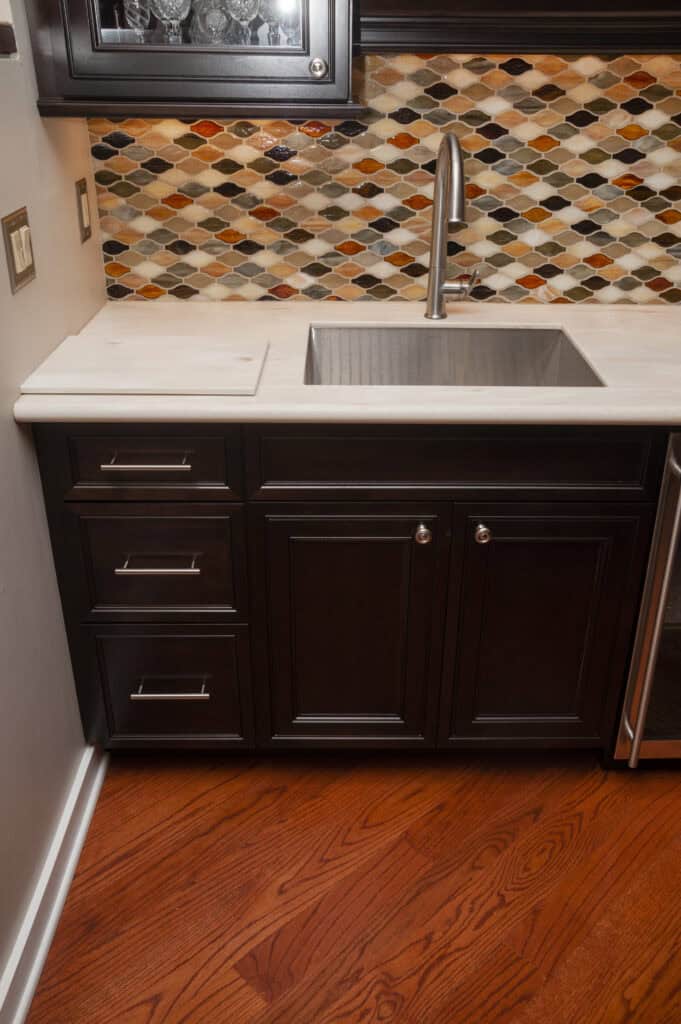 A kitchen sink with a modern faucet is set in a white countertop with dark wooden cabinets below. The backsplash features a colorful diamond tile pattern, and the floor is medium-toned wood.