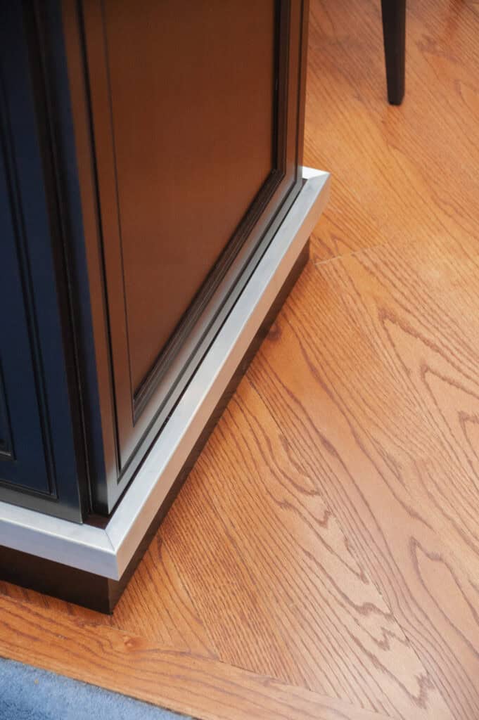 A close-up of the corner of a wooden cabinet with dark panels, featuring a metal trim at the base that sits on a reddish-brown hardwood floor.