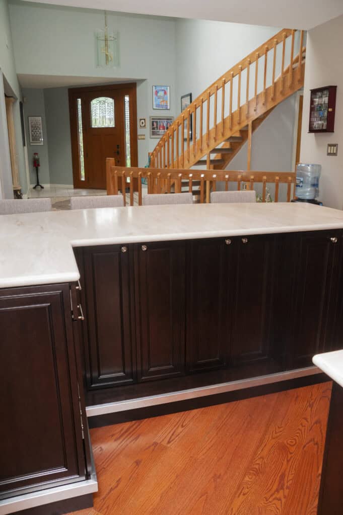A modern kitchen with dark wood cabinets and a marble countertop, overlooking an entryway with a wooden staircase and front door. The space features hardwood floors and light-colored walls.