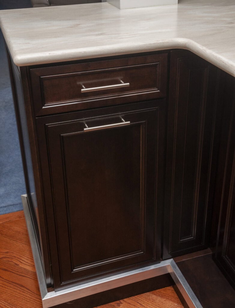 A close-up view of a dark wood kitchen cabinet with silver handles, positioned at the corner of a countertop with a beige marble surface and reddish-brown wood flooring.