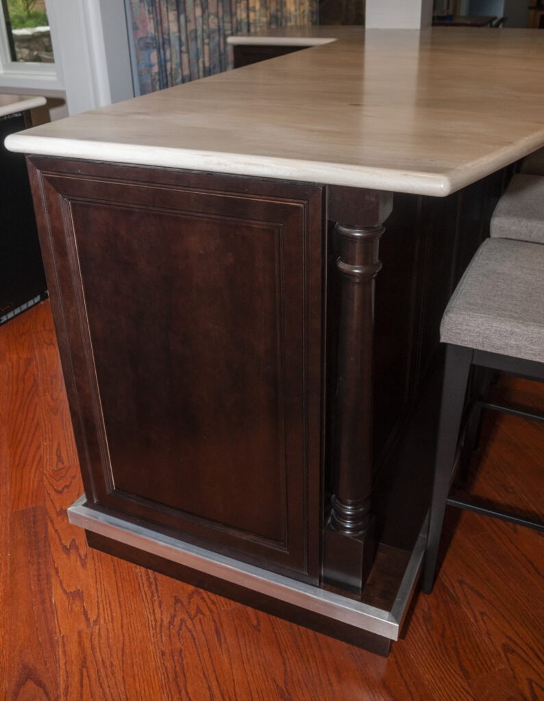 A dark wood kitchen island base with decorative trim and a light stone countertop, set on a reddish-brown hardwood floor next to upholstered bar stools.