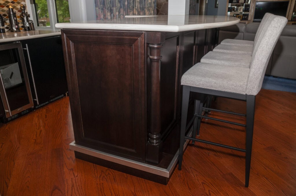 A kitchen island with a dark wood base and a light countertop, next to two upholstered barstools with light gray fabric seats and black metal legs, on a polished wooden floor.