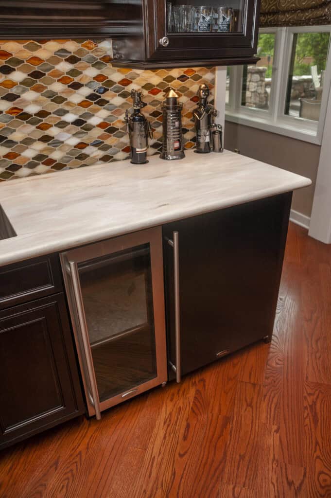 Modern kitchen with dark wood cabinets, a glass-front mini fridge, metallic bottle-shaped decor, colorful geometric tile backsplash, and medium-toned wood flooring. A window shows a stone patio outside.