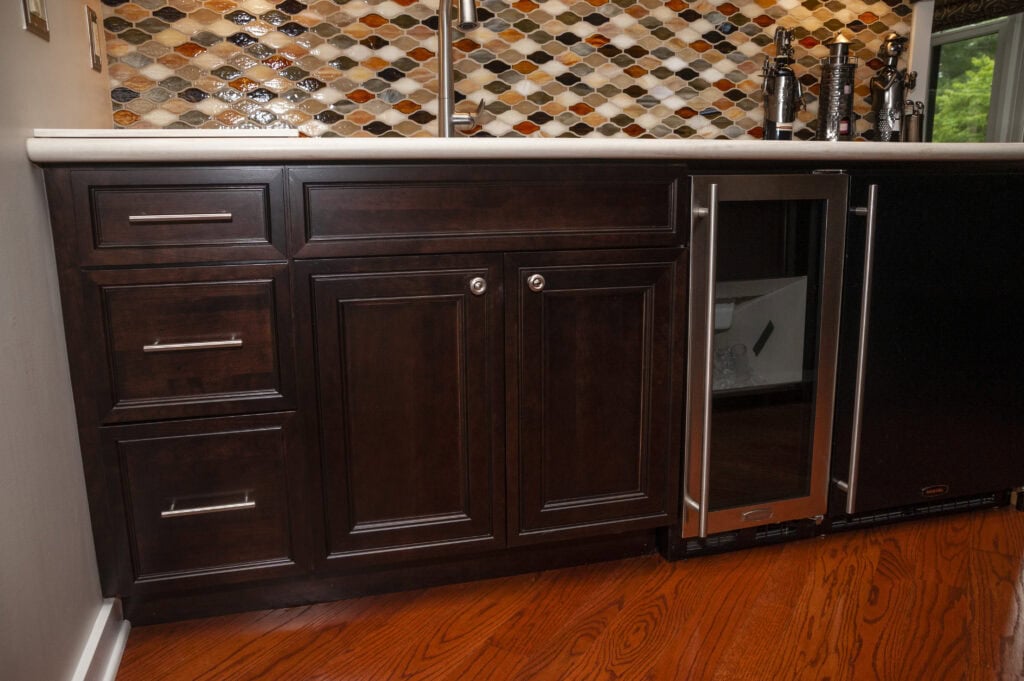 Dark wood kitchen cabinets with silver handles are set against a multicolored tile backsplash. The lower cabinets include drawers and a small glass-front refrigerator, all on a reddish-brown hardwood floor.