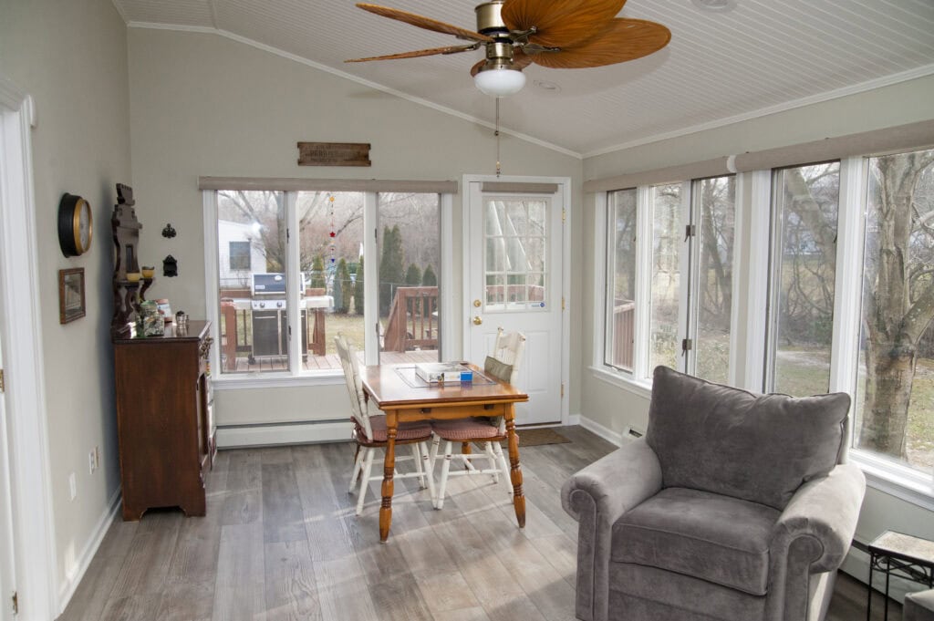 A bright sunroom with large windows, a wooden dining table with two chairs, a gray armchair, wood flooring, a ceiling fan, and a door leading out to a deck with trees visible outside.