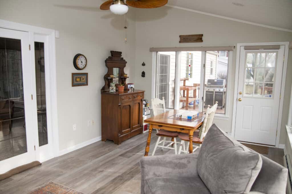 A cozy sunroom with a gray couch, wooden dining table with chairs, antique cabinet, wall clock, and glass doors leading outside. Sunlight streams in through windows and a glass door to a deck.