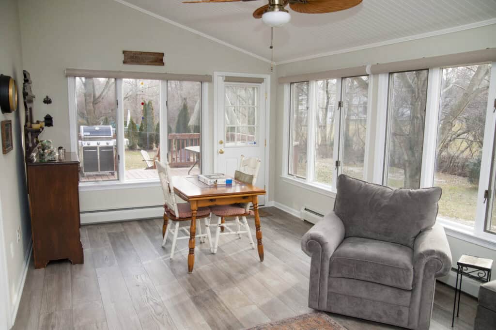 A bright sunroom with large windows, a small dining table with two chairs, a gray armchair, wood flooring, and a view of a deck with a grill and trees outside.