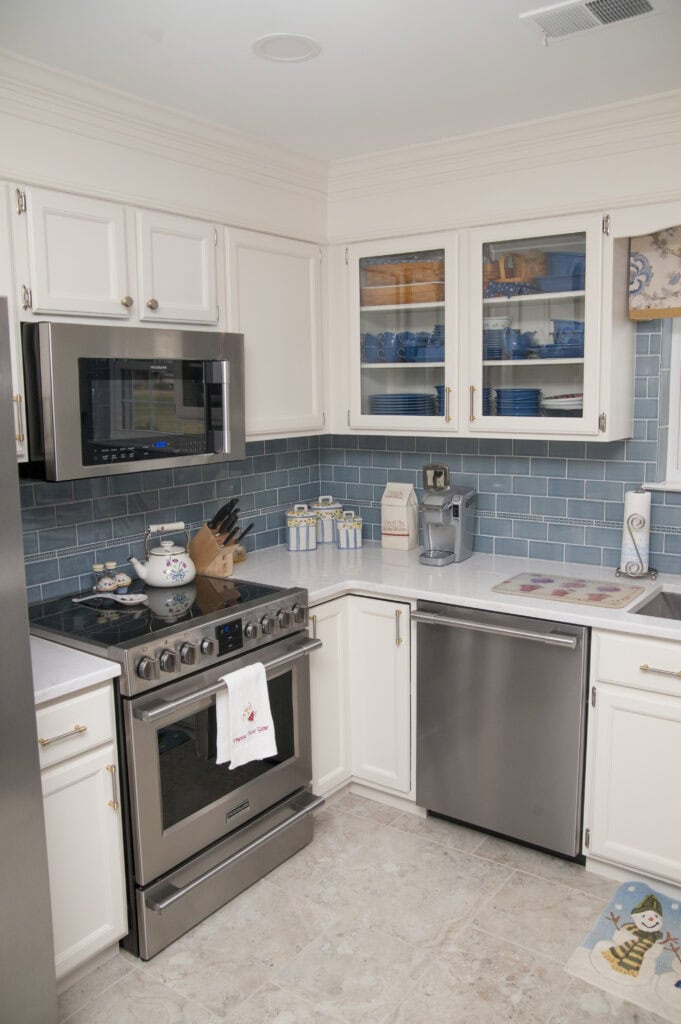 A modern kitchen features white cabinets, a stainless steel stove and dishwasher, blue subway tile backsplash, glass-front cabinets displaying dishes, and various small appliances on the countertops.