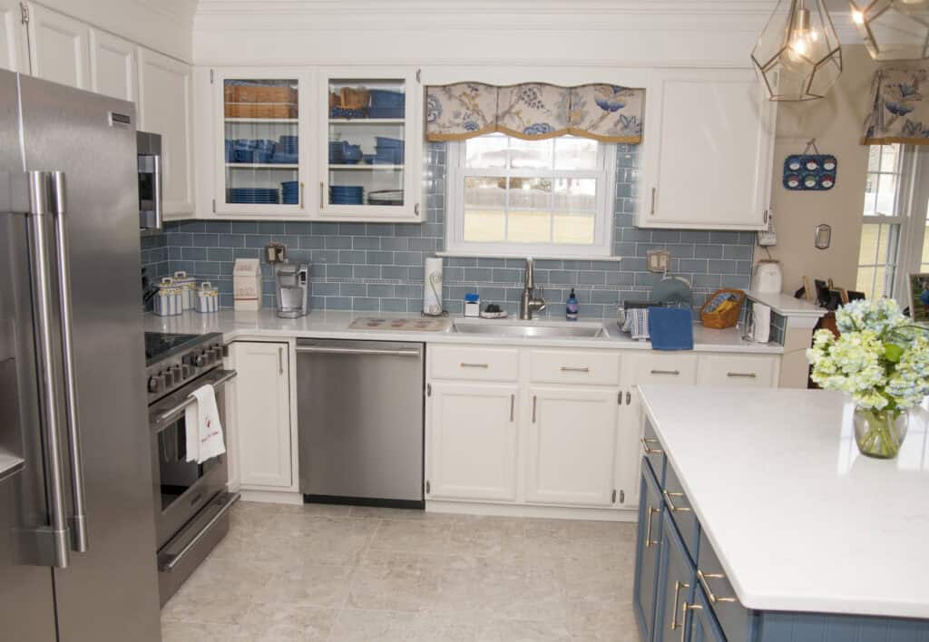 Modern kitchen with white cabinets, blue tile backsplash, stainless steel appliances, a window above the sink, and a white island countertop with a vase of flowers. Blue and white decor accents are visible throughout.