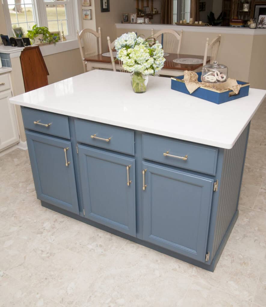 A kitchen island with blue cabinets, a white countertop, a vase of white flowers, and a decorative tray, set on a beige tiled floor in a bright room.