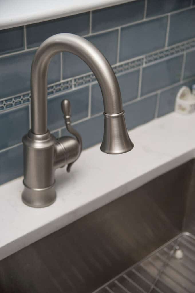 Close-up of a modern brushed metal kitchen faucet mounted on a white countertop, with blue subway tile backsplash and part of a stainless steel sink visible below.