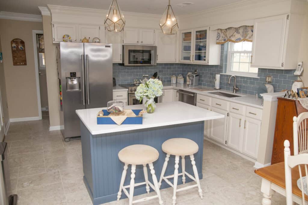 A bright kitchen with white cabinets, light blue tile backsplash, stainless steel appliances, a white island with two cream stools, and pendant lights. A vase of flowers and a tray are on the island.