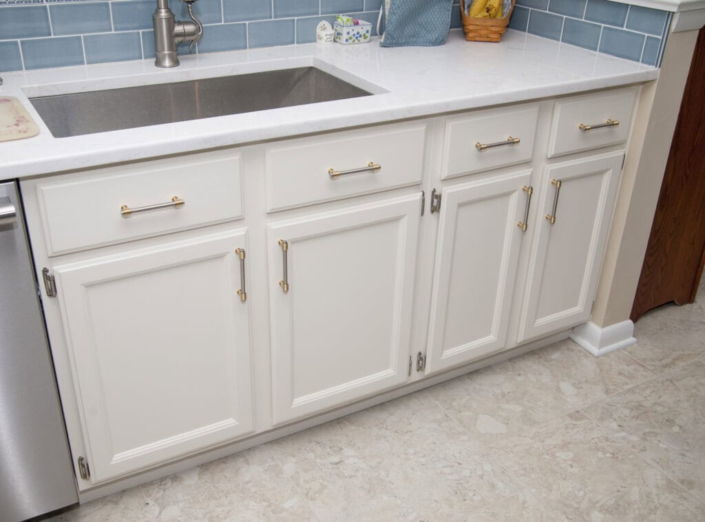 White kitchen cabinets with gold handles are beneath a white countertop and a stainless steel sink. The backsplash is blue tile, and there are items on the countertop, including a tissue box and a fruit basket.