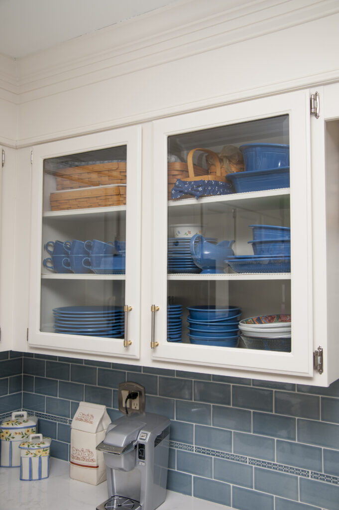 A white kitchen cabinet with glass doors displays blue dishes, bowls, mugs, and woven baskets on shelves. Below, a coffee maker sits on a counter with blue tile backsplash and containers for flour, sugar, and tea.
