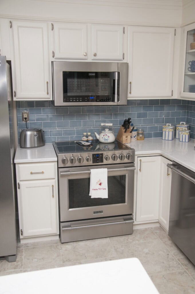 A modern kitchen with white cabinets, stainless steel appliances, blue subway tile backsplash, a stove with a towel, a microwave above, a toaster, and containers on the white countertops.