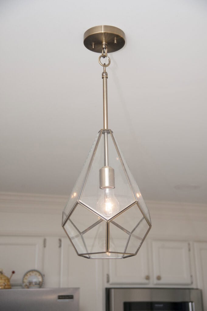 A modern pendant light with a geometric glass and brass frame hangs from a white ceiling, illuminating a kitchen with white cabinets in the background.